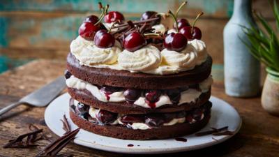 A Black Forest gâteau cake - with three layers of chocolate cake, cream and cherries - sits on a white plate on a wooden table 