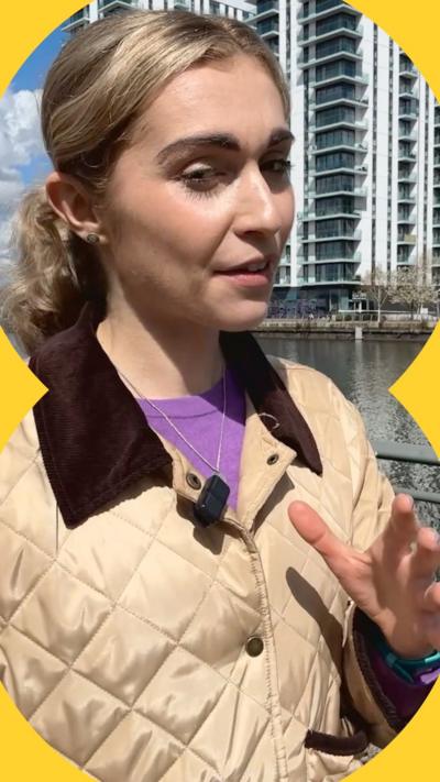 A woman with blonde hair wearing a beige jacket raises her left hand while talking.