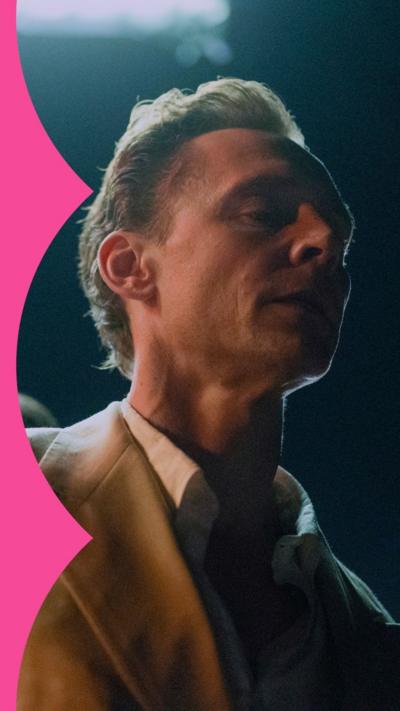 A man in a suit stands in the shadows looking worried