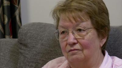 Sandra Holmes wearing a pink shirt and glasses. She is sitting on a grey sofa