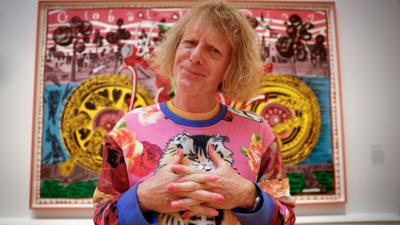 Grayson Perry folds his hands while posing in front of his painting