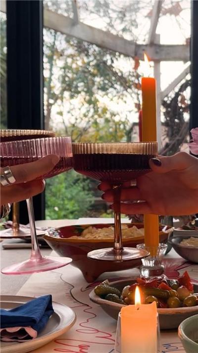 An orange candle sits next to bowls of food on a table Plates with napkins on can be seen in the background. The candle is lit. Two people are clinking their glasses together in the foreground.