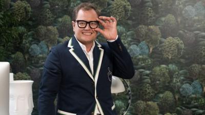 Alan Carr fixes his glasses as he smiles to the camera