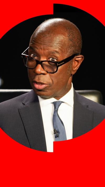 A man wearing a suit and glasses talks to the camera
