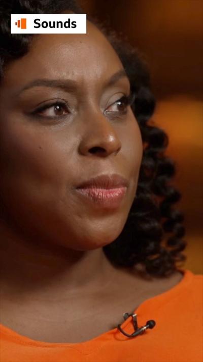 Author Chimamanda Ngozi Adichie looks to the side - she has curly black hair and wears an orange top