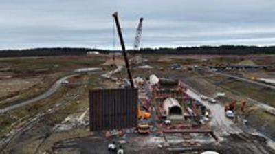 The image is taken from the air by a drone.  It shows a large construction site, where a new reservoir is being built.  There is a crane in the background, and another crane in the foreground, which is in the process of lifting a large brown steel panel, which is a new wall at the reservoir.  To the right there is a concrete tunnel and a trench where the wall will be lowered into place.