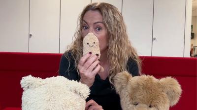 Women with blonde, wavy hair is sitting on a red sofa with teddy bears in front of people.