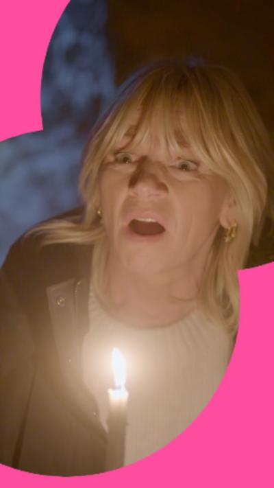 Zoe Ball has long blonde hair she is wearing a white top and a dark coat by candle-light. She looks shocked.