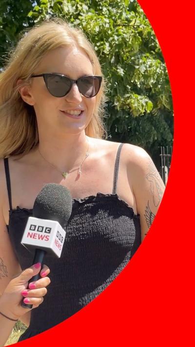 A woman speaking into a mic. She has blonde hair, sunglasses, a black strappy dress and tattoos.