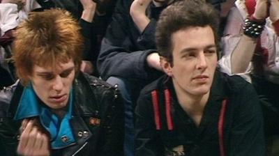 Paul Siminon and Joe Strummer of The Clash sitting being interviewed in a studio, with an audience behind them. Paul on the left with light red dyed hair, in a light blue shirt under a black leather jacket. Joe on the right, with dark hair and wearing a black shirt with red stripes on it.