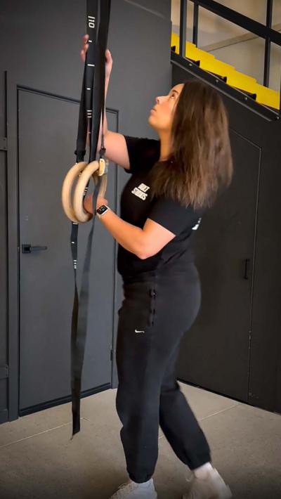 A woman with brown hair and wearing black t shirt and tracksuit bottoms moves gym equipment