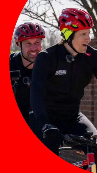 Greg James at the front of a tandem bike with Prince William sitting behind him. They both smile and wear black long-sleeve tops and red helmets.