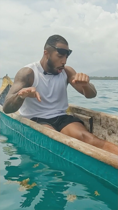Race Across The World contestant Tyler West in a narrow fishing boat looking shocked after catching a fish. He's holding a fishing line in his left hand and moving his right hand away and wearing sunglasses on his head.