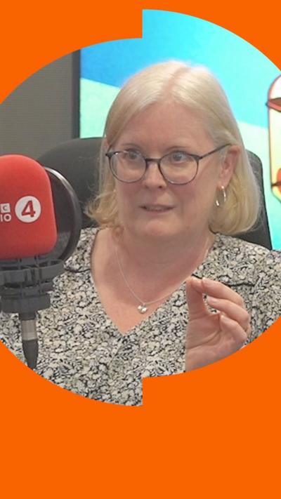 Woman in patterned top with shoulder length blonde hair and glasses sits in front of a microphone