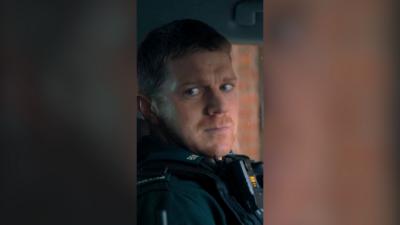 A uniformed police officer in a car, looking wearily to his right. He has short ginger hair and stubble