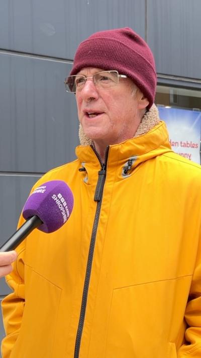 a man in a yellow coat and claret hat and glasses talking to a reporter