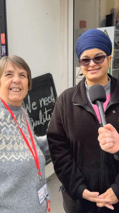 Two women smile at the camera. Woman on left wearing a grey jumper, red lanyard and brown hair. Woman on right is wearing a hijab, glasses and a black fleece cardigan. A microphone is held in front of them.