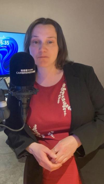 Female reporter stands in BBC Radio Cambridgeshire studio next to a microphone