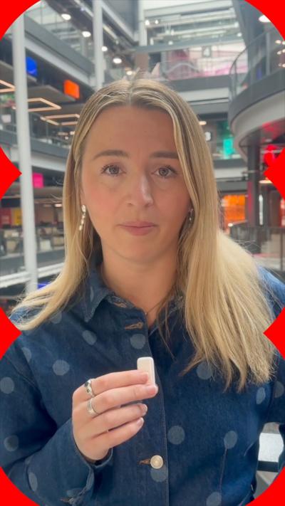 Lucy Woodham looks at the camera and holds a microphone with her right hand. She has long blonde hair and wears a denim dress with spots.