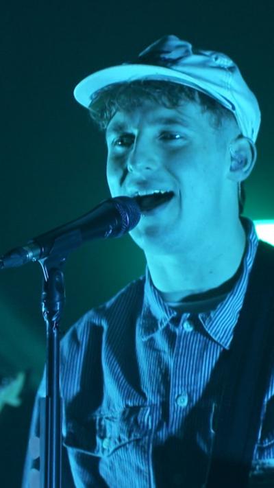 The Snuts perform live on stage. Jack the vocalist/guitarist is clearly visible as he stands in front wearing a light cap, a light jacket and jeans. The other band members are standing behind and aren't so clearly visible. The lights on stage are turquoise.