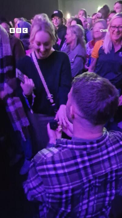 A man on bended knee proposes to a woman at a concert