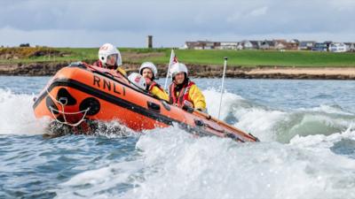 A three-person RNLI crew in yellow, wearing helmets, on board a yellow lifeboat on the sea.