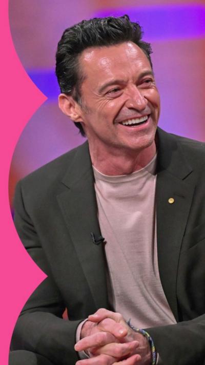 Hugh Jackman smiling in Graham Norton studio. He wears a grey suit with pale grey t-shirt underneath.