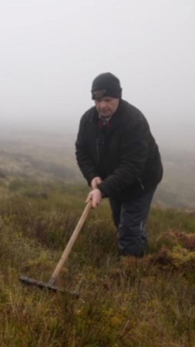 A man in a black jacket and black hat is using a rake to pull moss out of the ground, he is also wearing dark trousers. In the background there is moss covering the entire ground in various colours including green, brown and yellow, the sky is full with fog.