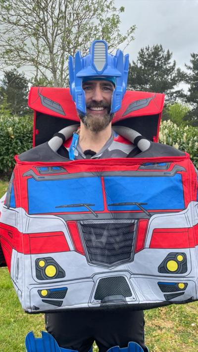 A man stands smiling wearing an Optimus Prime costume outside with trees and grass behind him