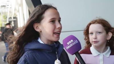 A young girl with long brown hair wearing a coat with a rugby ball keyring on the zip speaking into a Newsround mic