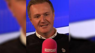 Man in a dark jumper and white shirt sitting behind a red radio microphone with Scotcast on it.
