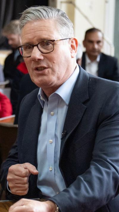 Starmer holds his hand in a fist and has a neutral expression as he speaks while sat at a table
