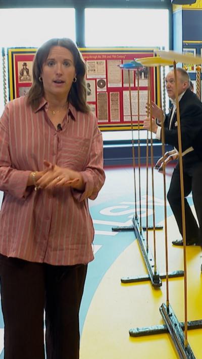 Woman in pink top and black trousers next to circus performer in black tuxedo spinning plates