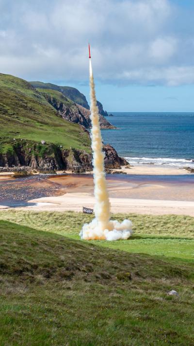 Rocket launches from grass-covered land near a beach by the sea