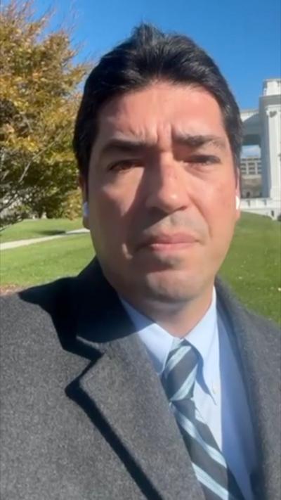 Bernd Debusmann wearing grey coat, blue shir and blue & dark grey tie looking into phone camera.