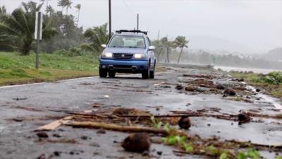 A blue car drives along a road littered with branches next to the sea