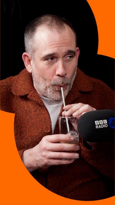 Xand van Tulleken blowing into a glass of water with a straw. He's wearing an orange jacket.