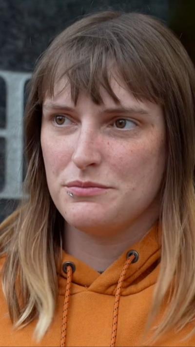 A woman in an orange hoodie looks to the side as she speaks to the camera