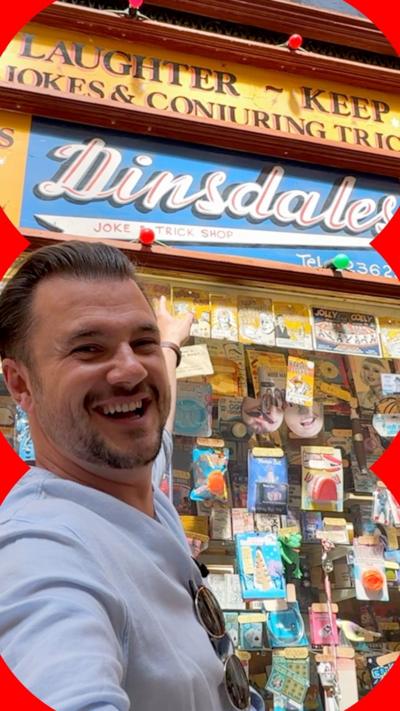 A BBC journalist standing in front of the Dinsdales joke shop and smiling