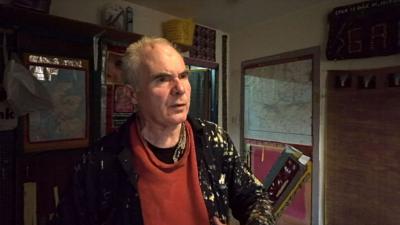 Dan MacNee standing in his cluttered flat in a red jumper and black jacket spattered with white and yellow paint.