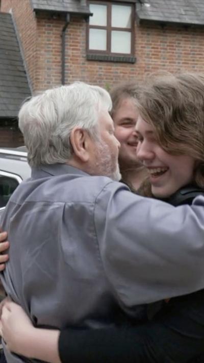 A man wearing a grey shift with grey hair hugging two young men with long hair. They are all smiling