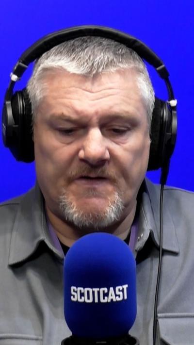 John Davidson sits in front of a radio studio microphone, he wears headphones and a grey shirt