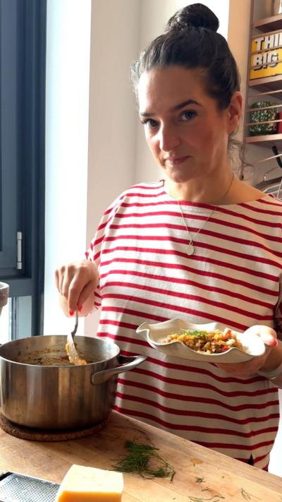 A woman wearing a white and red striped top looks at the camera whilst holding a plate with one hand and dipping a spoon in a pot with the other.