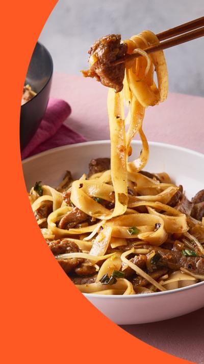 A pair of chopsticks holds noodles from a bowl of beef chow fun