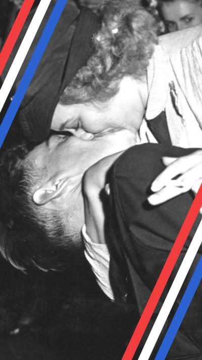 A close-up black and white photo of a woman with curly hair wearing a naval cap. She has a cigarette between her fingers and is holding a uniformed sailor below her, kissing him passionately on the lips during the VJ Day celebrations in the middle of a crowded New York City.