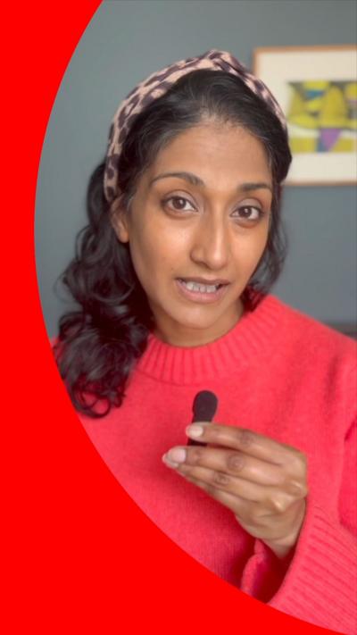 A woman wearing a red jumper holds a small microphone and looks directly at the camera