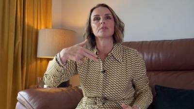 Rachel Williams sitting on a brown leather sofa while wearing a yellow, black and white dress. Her hand is making a gun gesture