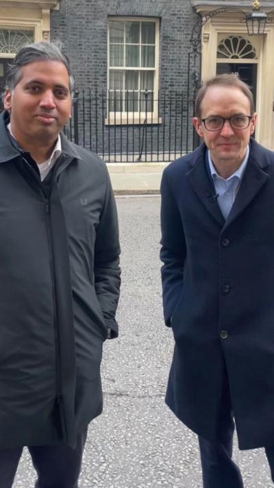 Two male journalists stand next to each other on Downing Street. One wears a dark green jacket and the other wears glasses and a navy jacket