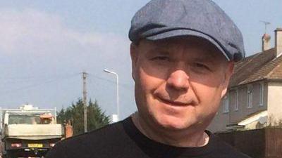 A head and shoulders photo of a man wearing a grey hat smiling at the camera with a residential street behind him.