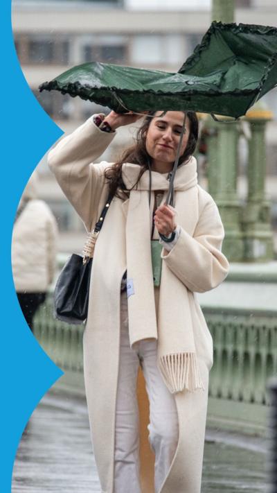 A woman with long brown is struggling with a green umbrella. She is wearing white trousers, a long cream coat and cream scarf, and a carrying a large, dark-coloured handbag.